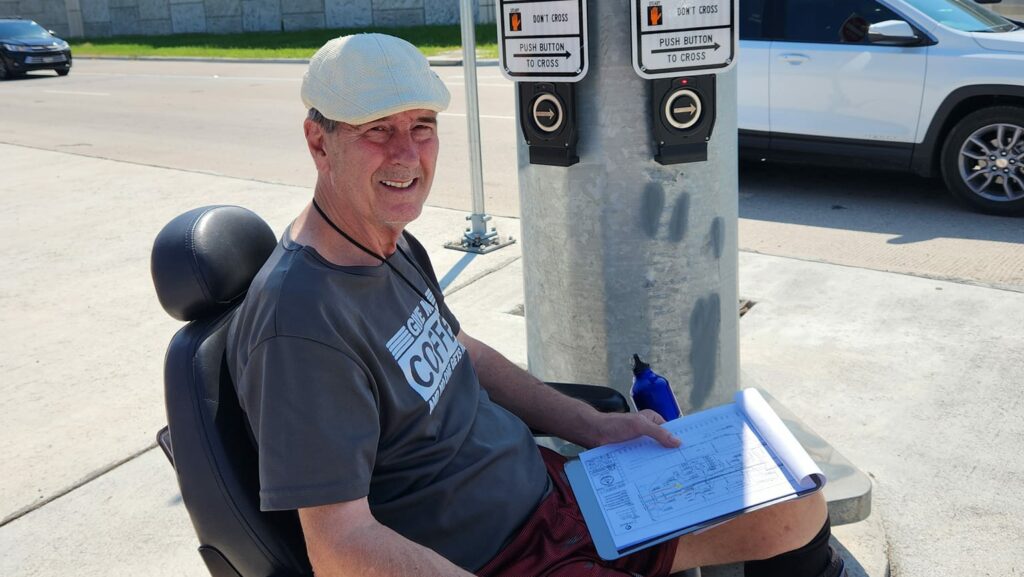 White man in a motorized wheelchair sitting next to a pedestrian crossing signal and holding a clipboard.