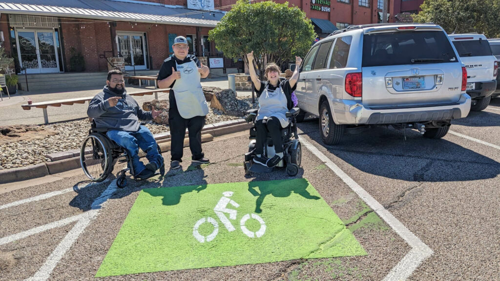 A man and a woman in wheelchairs and a second man standing lift their arms in celebration. They are in a parking space with green and white markings indicating that this spot is for bicycle parking.