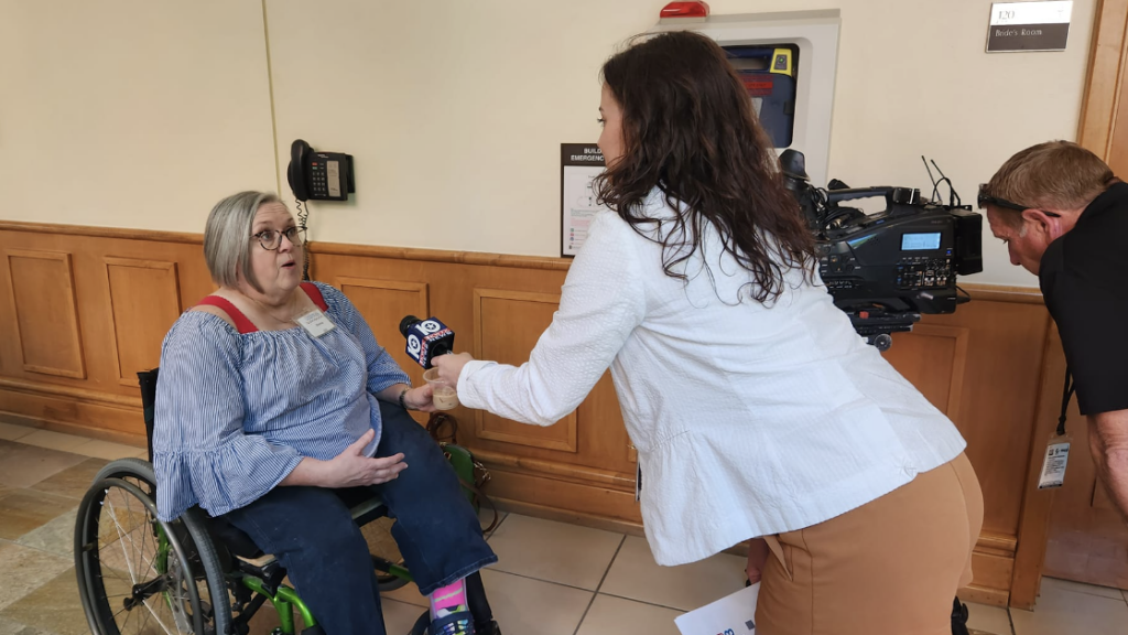 A white woman wearing glasses and sitting in a wheelchair being interviewed by a white woman holding a microphone. A white man looking in a television camera is on the right edge of the frame.