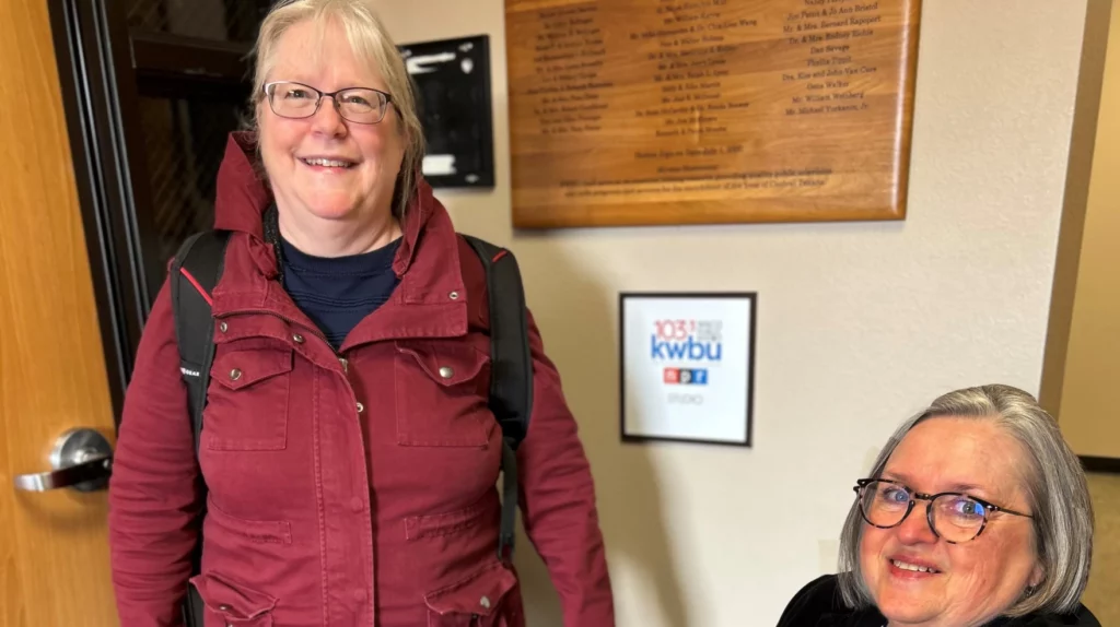 One woman standing and another sitting, both with gray hair and wearing glasses, with a sign between them that reads 103.3 Waco Public Radio KWBU NPR