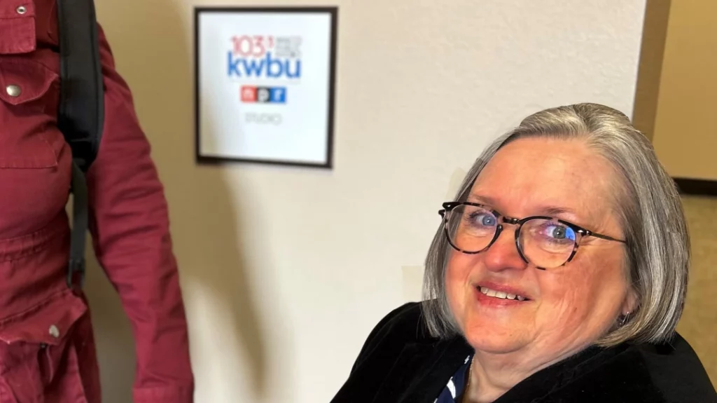 A woman with gray hair, wearing glasses, is sitting next to a sign that says 103.3 Waco Public Radio KWBU NPR