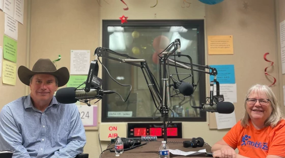 Two people sitting across a table from one another in a radio studio. The man on the left is wearing a cowboy hat. The woman on the right is wearing a bright orange T-shirt with the Mobilize Waco logo