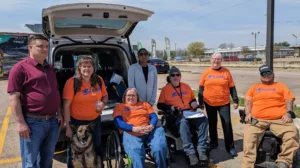 7 people, 4 wearing orange Mobilize Waco T-shirts, 3 in wheelchairs, one with a Seeing Eye dog, posing in front of the tailgate of a van, some holding clipboards