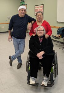 Two women and a man. One of the women is in a wheelchair. The two people standing behind her are dressed for a Christmas event.