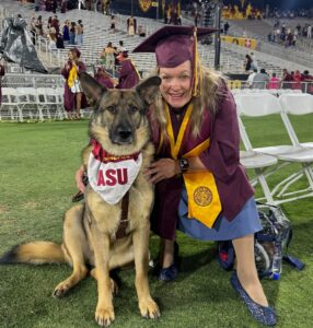 A woman wearing a maroon graduation cap and gown, accompanied by a German shepherd Seeing Eye dog wearing a scarf that says ASU.