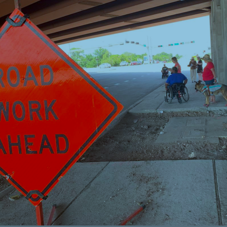 An orange sign reading Road Work Ahead is in the foreground. In the background are six people standing on the other side of a badly damaged sidewalk. One of the people has a seeing eye dog. Two other people are sitting in wheelchairs.